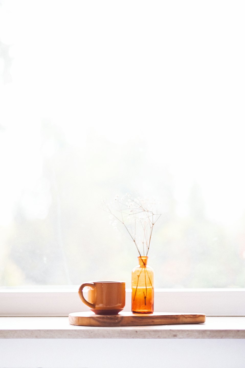 Taza y flores secas sobre una bandeja de madera junto a una ventana.